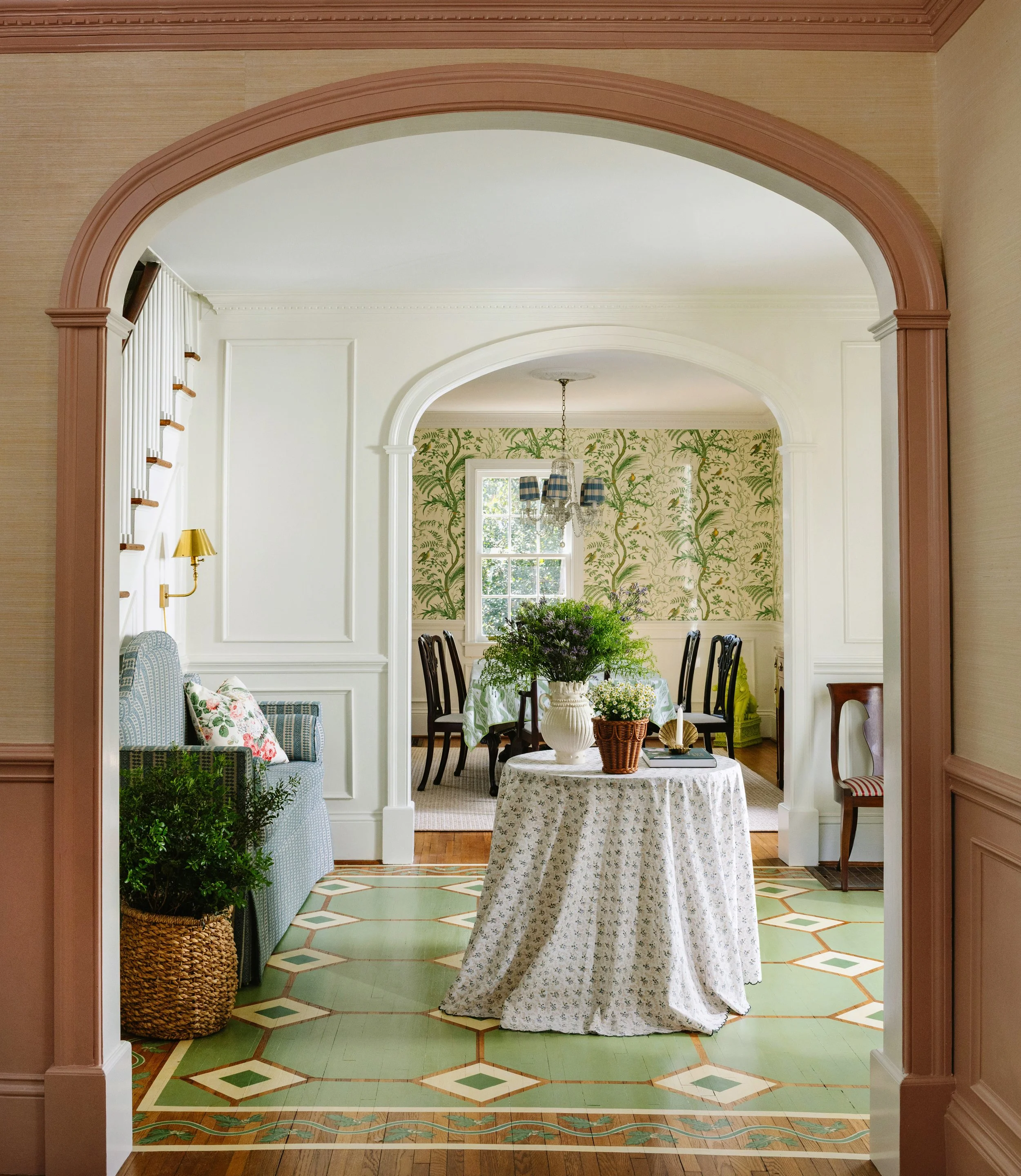 View of a bright dining area with a round table covered with a floral tablecloth and a large vase of greenery on top, seen through an arched doorway from an adjacent room with patterned green and cream wallpaper.