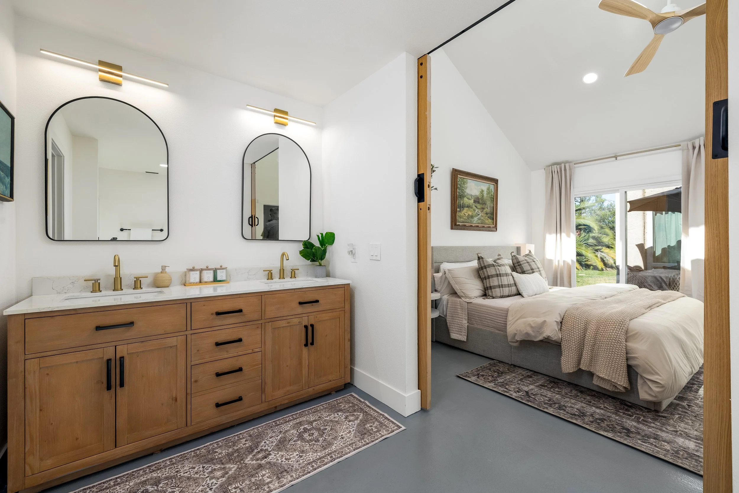 Modern bathroom with wooden double vanity, arched mirrors, and gold fixtures adjoining a bedroom with neutral decor and large window view.