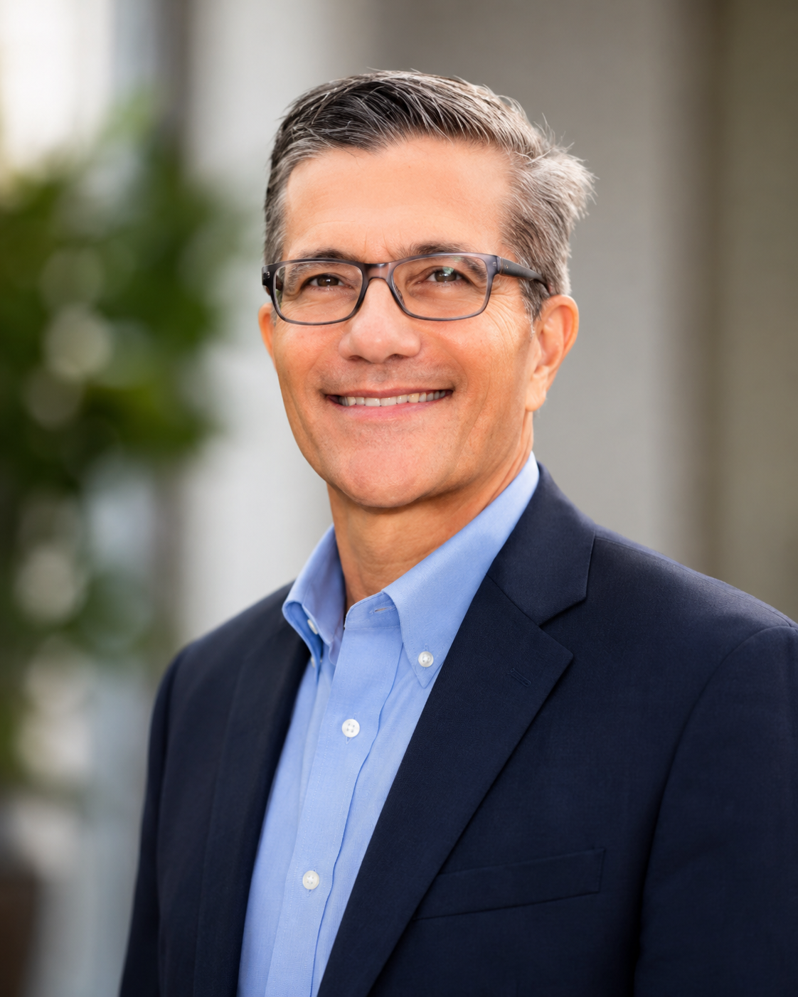 A smiling middle-aged man wearing glasses and a blue suit jacket, standing outdoors near a body of water with trees and greenery in the background.