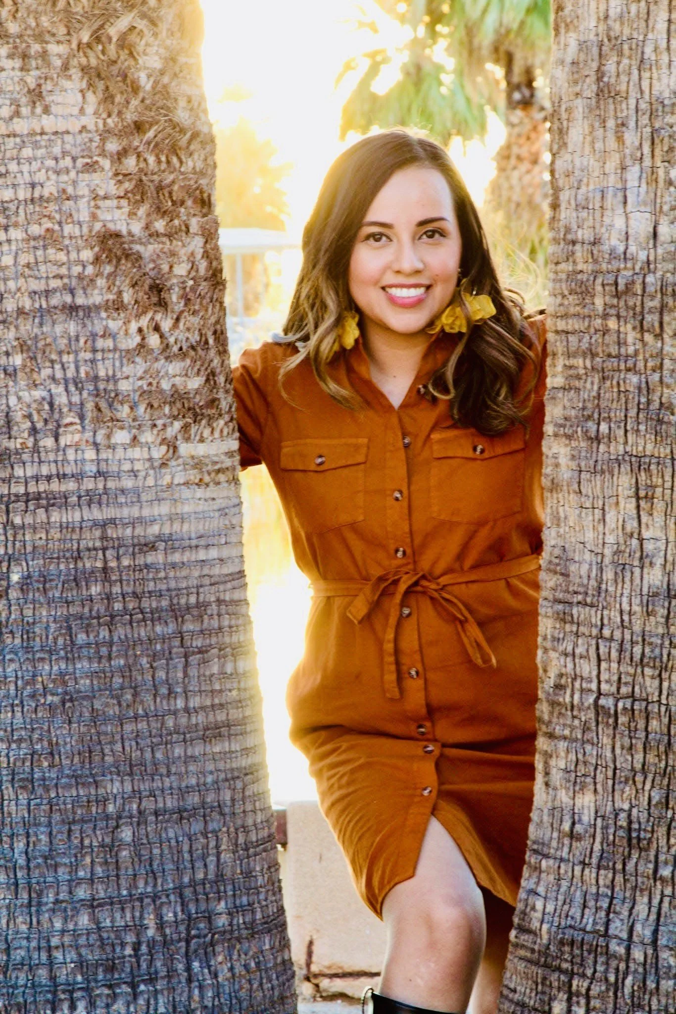 A woman smiling in a brown dress with a belt, standing between two tall palm trees at sunset.