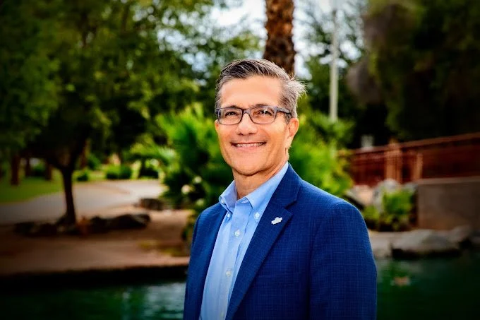 A smiling middle-aged man wearing glasses and a blue suit jacket, standing outdoors near a body of water with trees and greenery in the background.