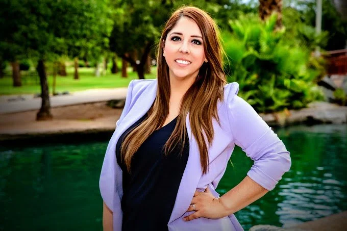 A woman with long brown hair in a black top and light purple blazer standing outdoors near water and trees, smiling at the camera.