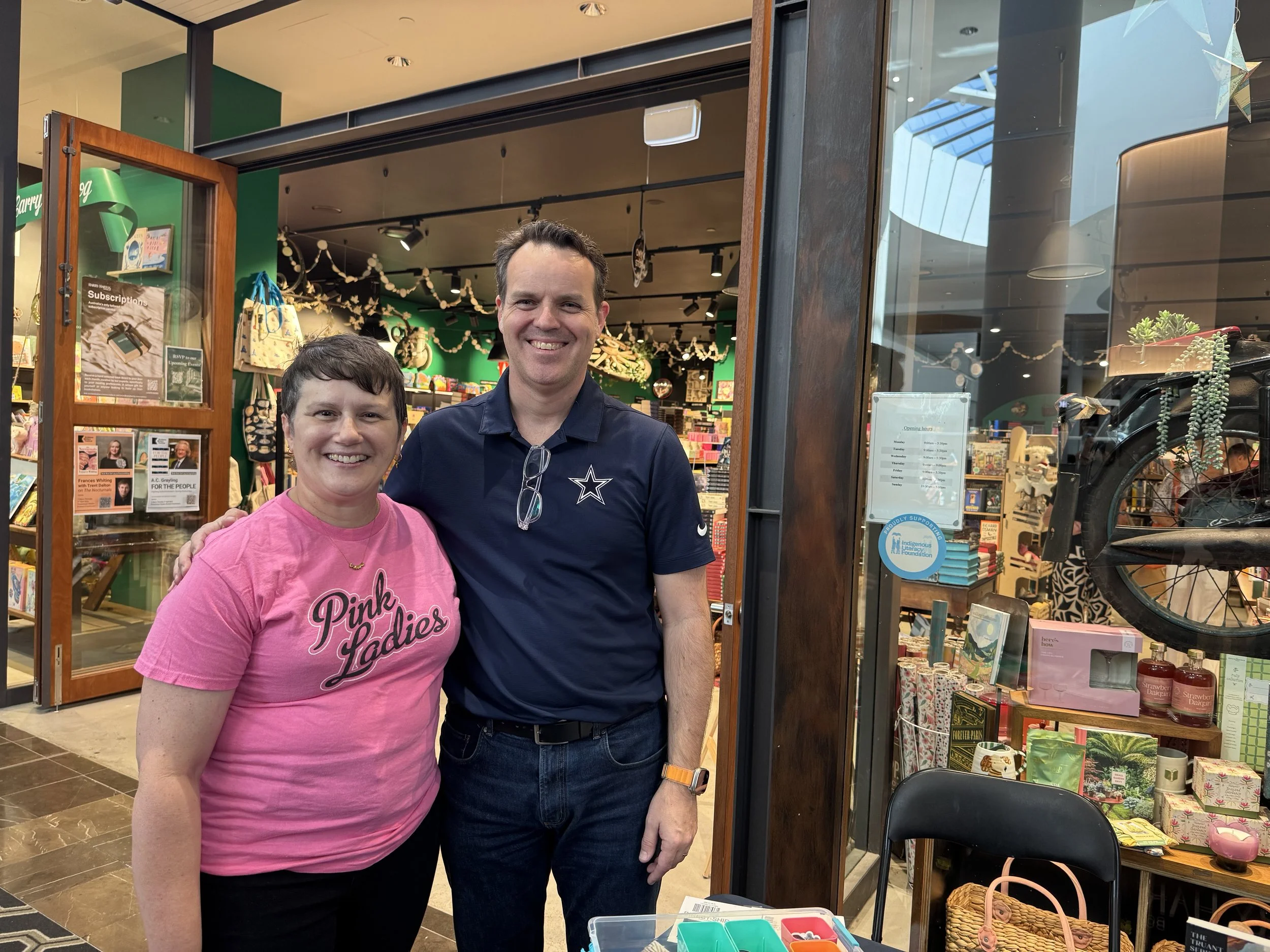 A smiling woman and man standing together inside a store, with shelves of colorful items and decorations behind them.