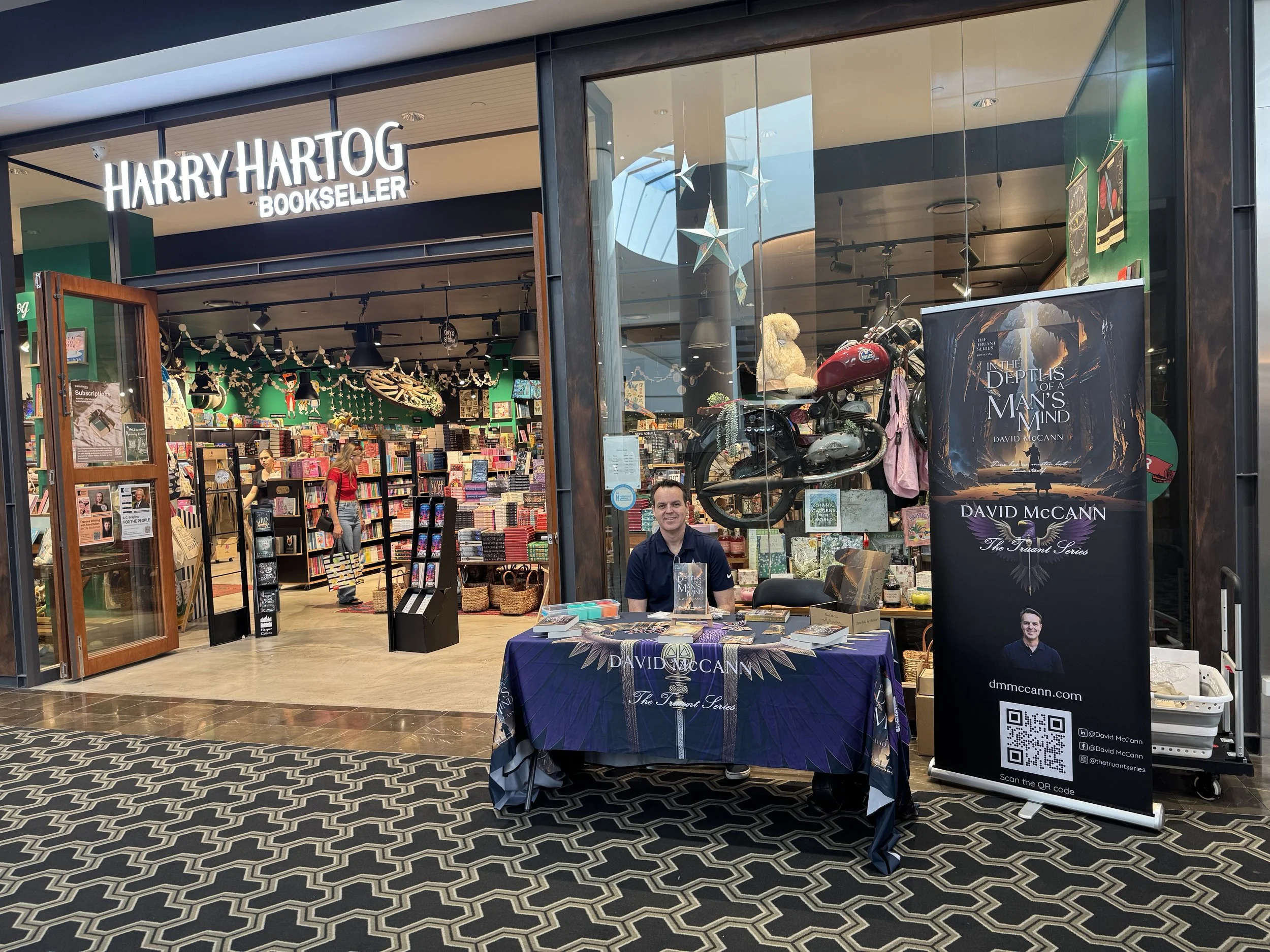 Author David McCann is seated at a table outside Harry Hartog Bookseller, promoting his book "In the Depths of a Man's Mind." The table is draped with a dark cloth displaying his name and book series. A large promotional banner with the book cover and author photo stands next to the table. The store's interior is visible through the open door, with shelves of books and festive decorations.