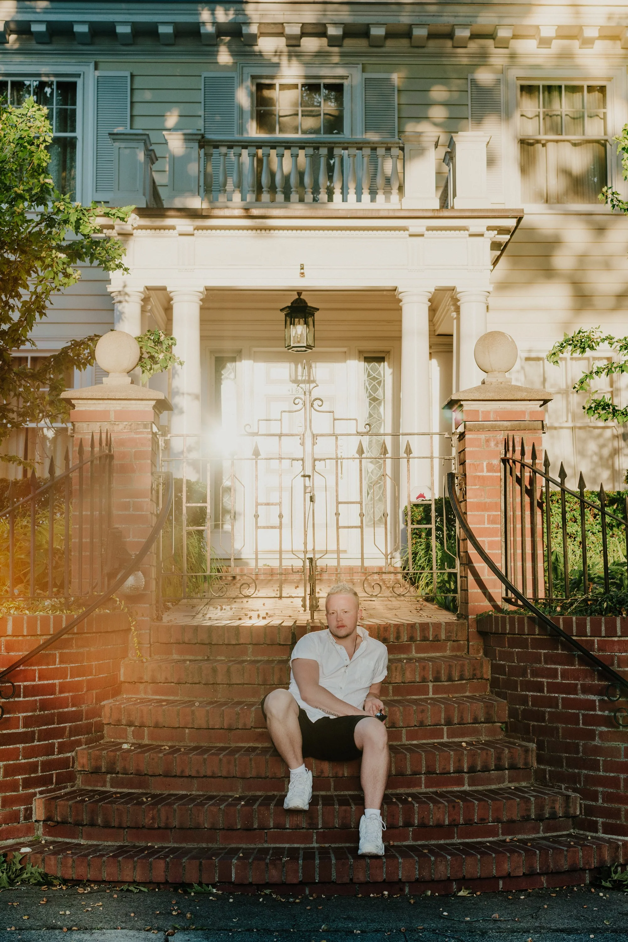 A young man in a white shirt and black shorts sitting on brick stairs in front of a large house with a gate and columns, with sunlight filtering through trees.
