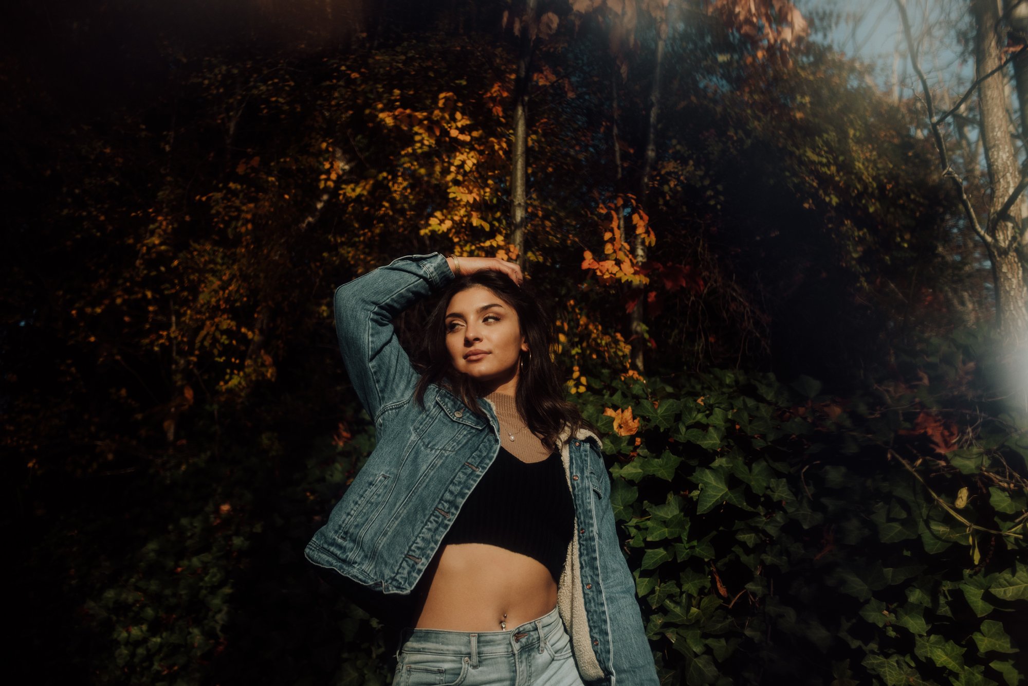 A young woman standing outdoors in front of foliage during autumn, wearing a denim jacket, crop top, and jeans, with one hand on her head and looking away.