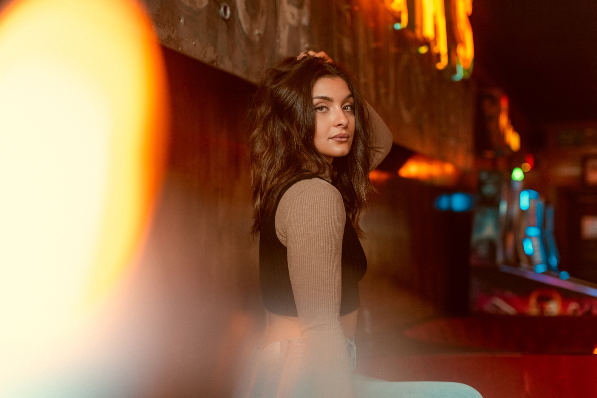 A young woman with wavy dark hair and light skin poses in a dimly lit bar or arcade with colorful neon lights in the background.