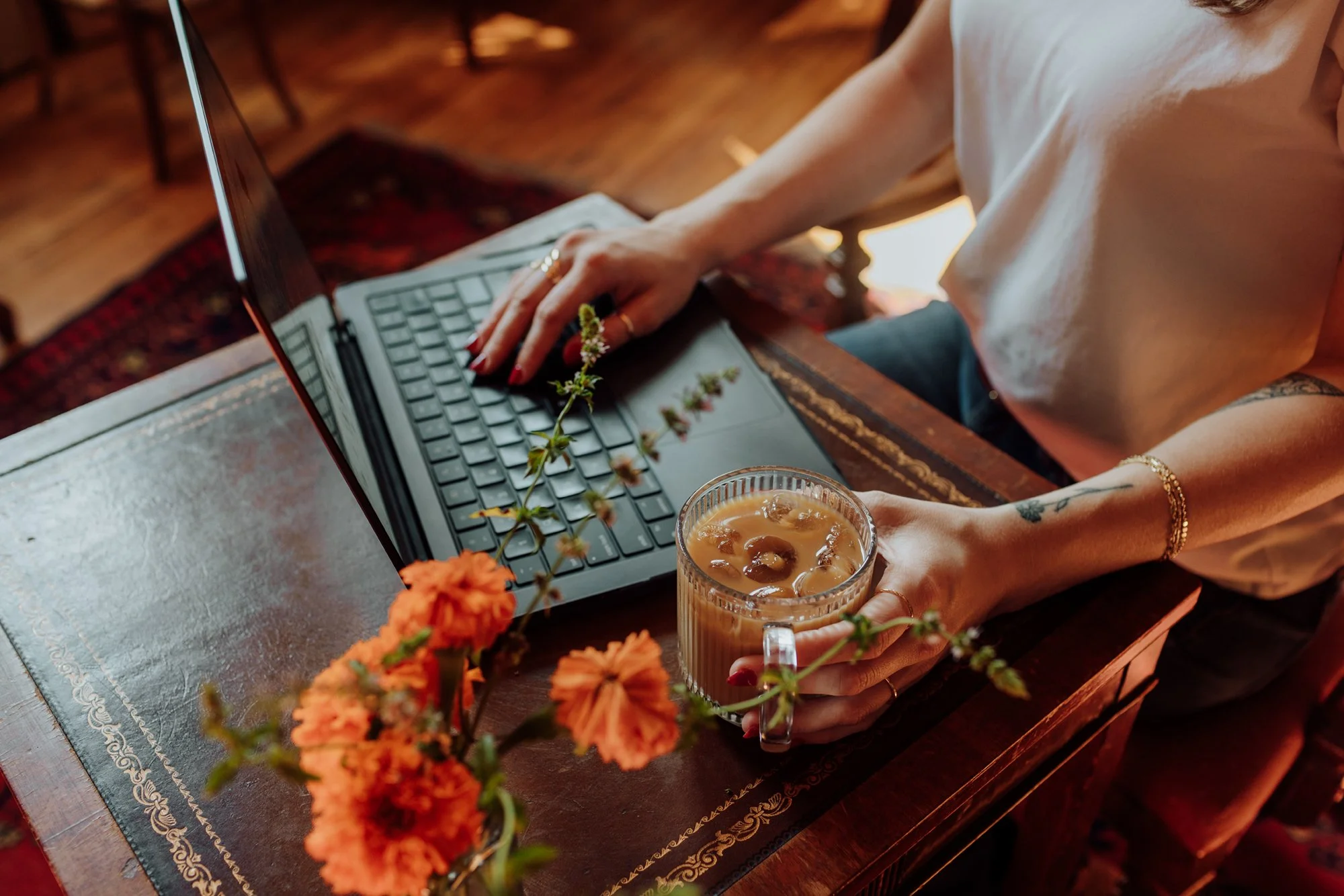 Person working on a laptop at a wooden table with a floral arrangement and a glass of iced coffee.