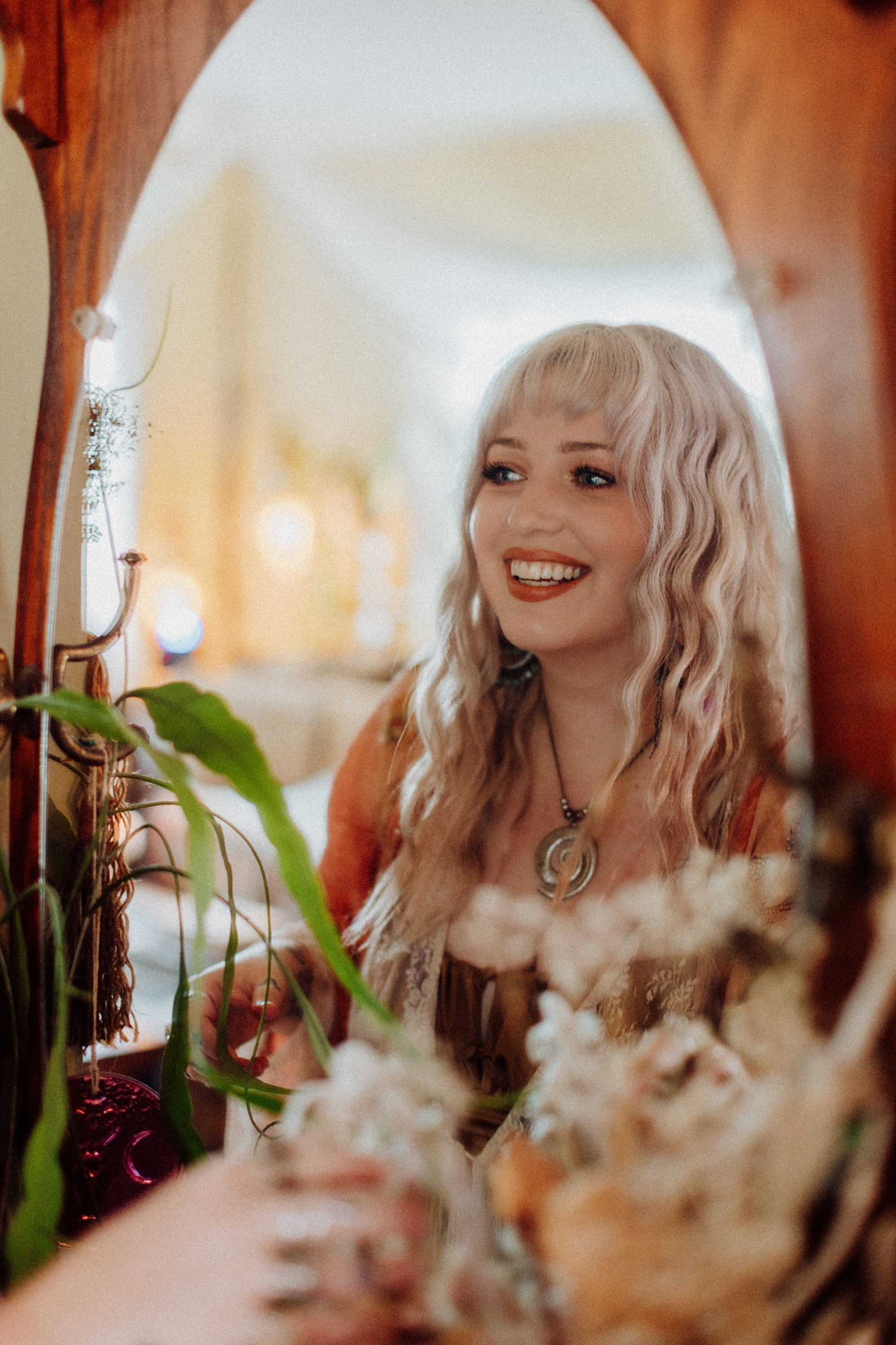 A young woman with curly blonde hair smiling while looking into a mirror surrounded by plants and flowers.