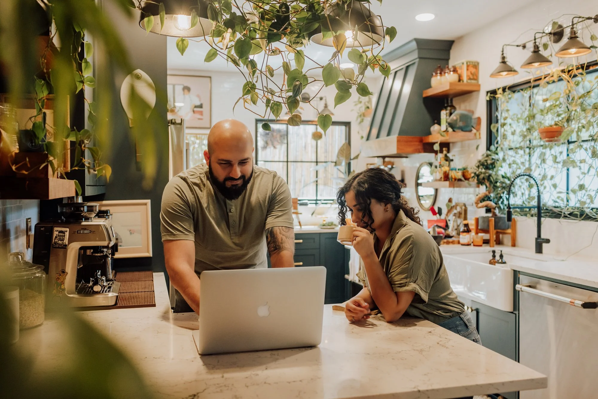 A man and woman in a modern kitchen looking at a laptop on the counter, woman drinking from a mug, surrounded by greenery and kitchen shelves.