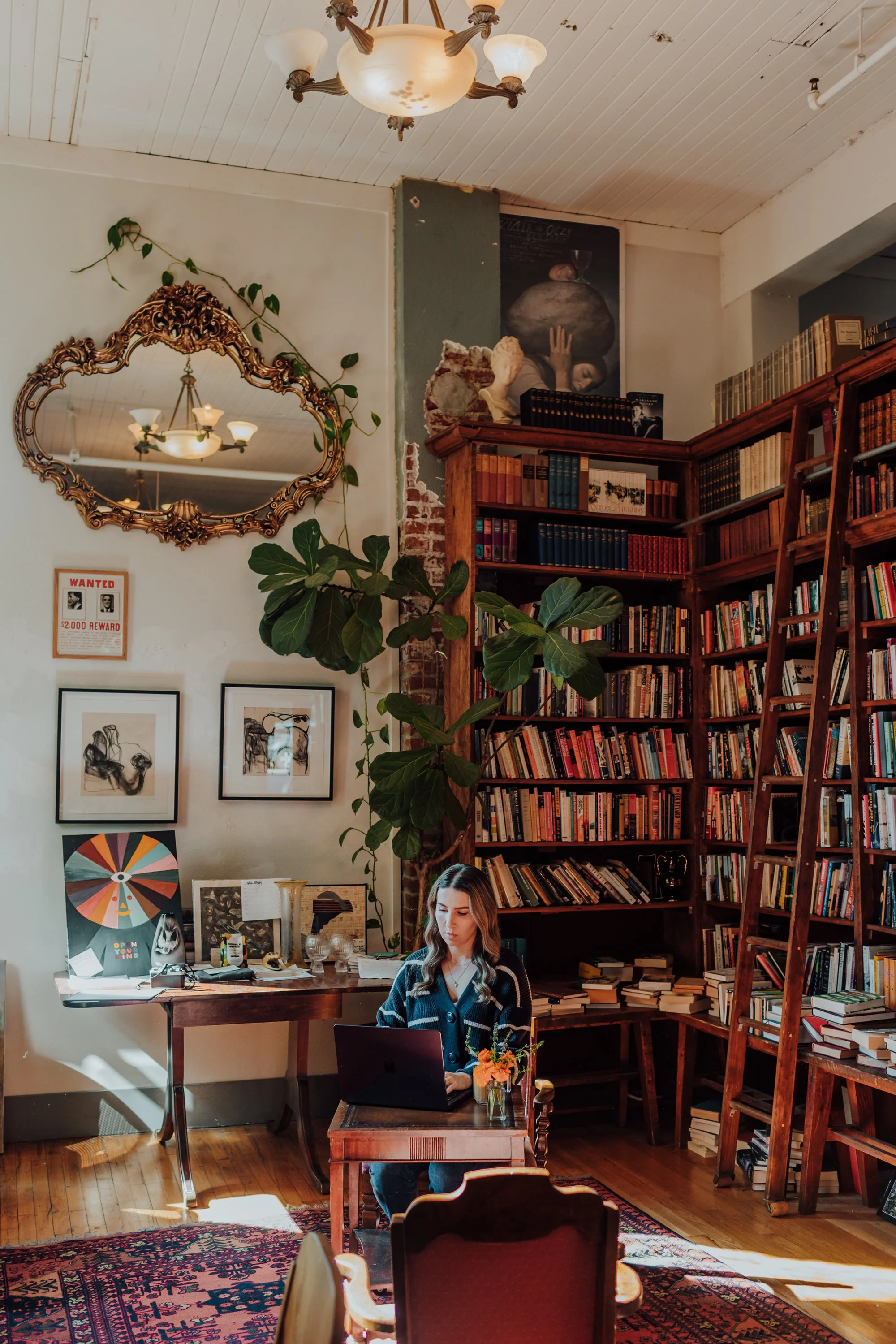 A woman working on a laptop at a small wooden table in a cozy library or study room filled with bookshelves and artwork, with sunlight streaming through a window.