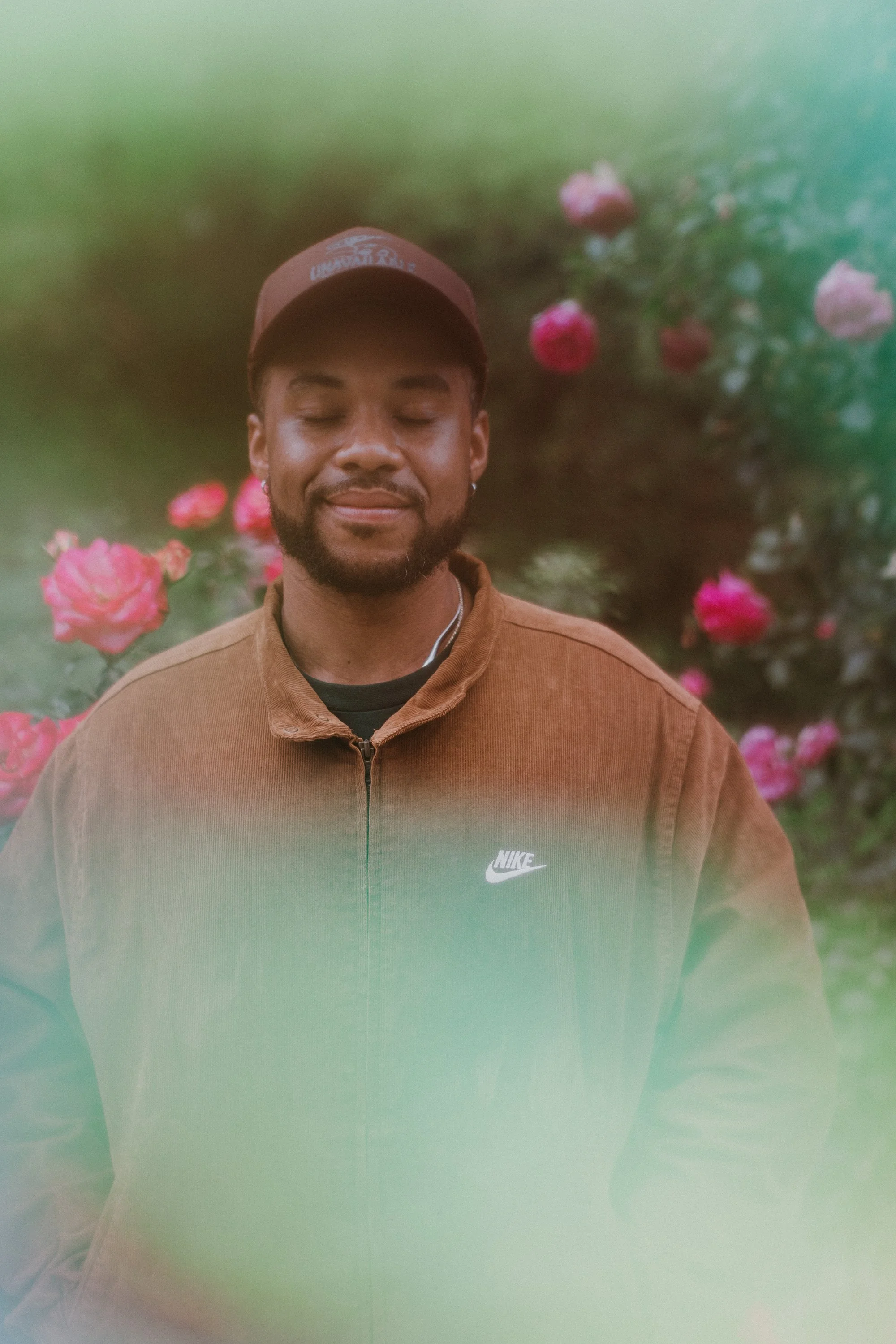 A man with closed eyes and a slight smile, wearing a brown Nike jacket and a brown cap, standing amidst pink roses with a greenish-blurred background.