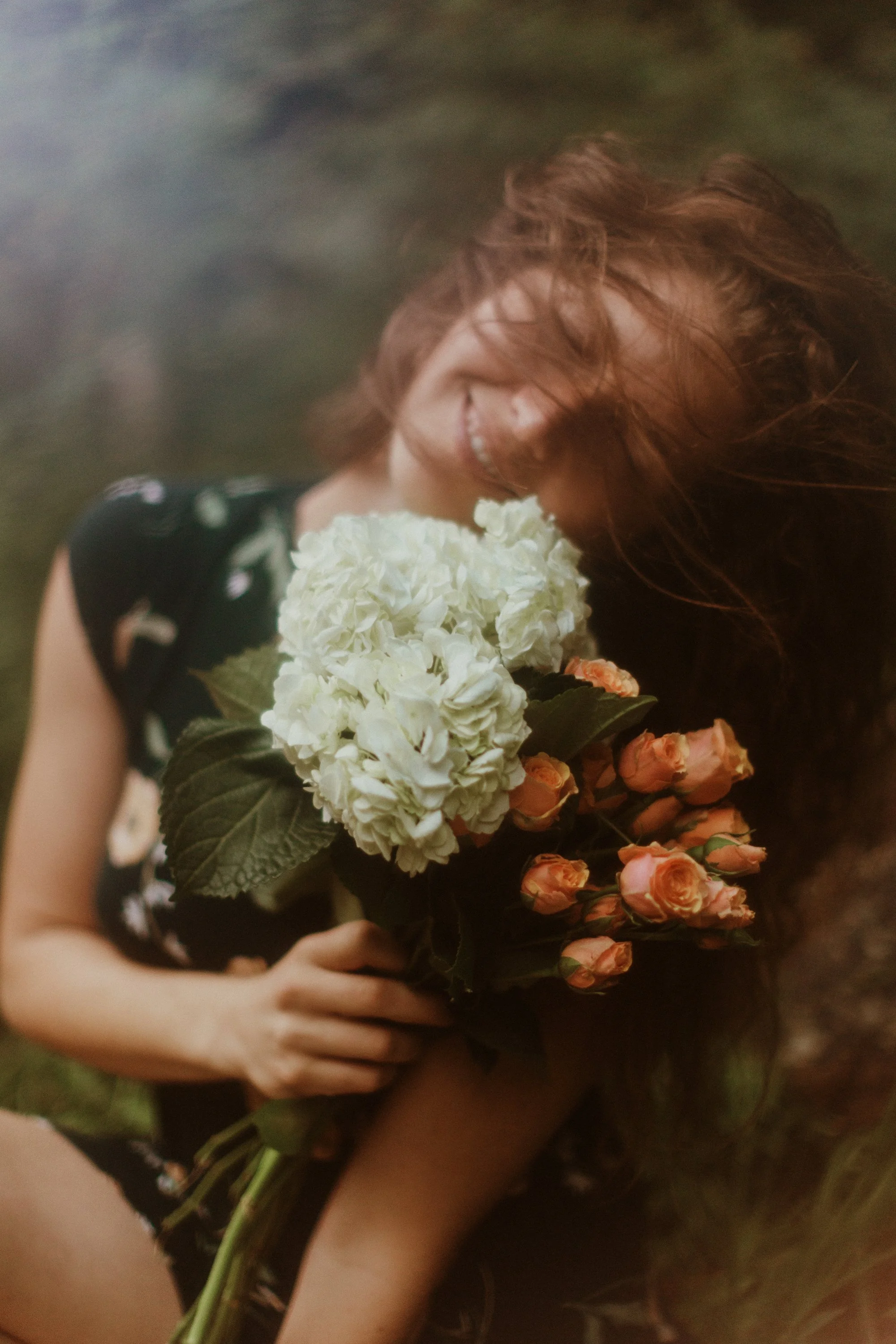 A woman with long red hair smiling and holding a bouquet of white and pink flowers outdoors.