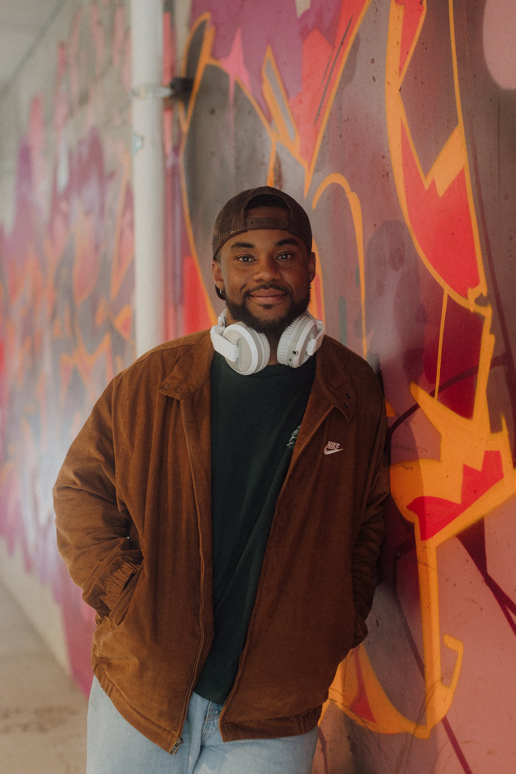 A man standing in front of a colorful graffiti wall, wearing a brown jacket, black Nike shirt, baseball cap, and white headphones around his neck.