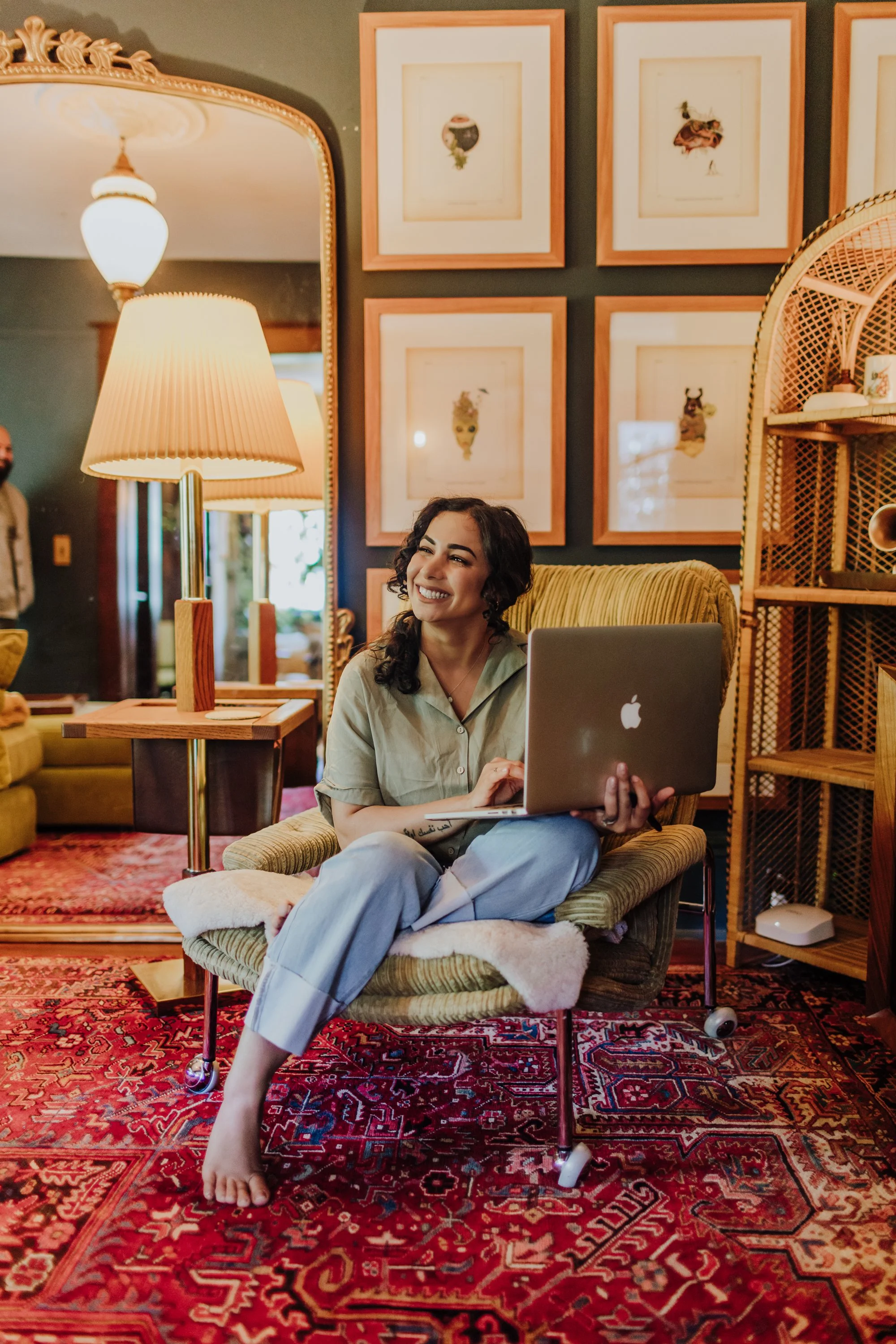 A woman sitting on a green armchair, smiling while holding a silver MacBook laptop, in a cozy room with framed artwork on the wall and a red patterned rug.