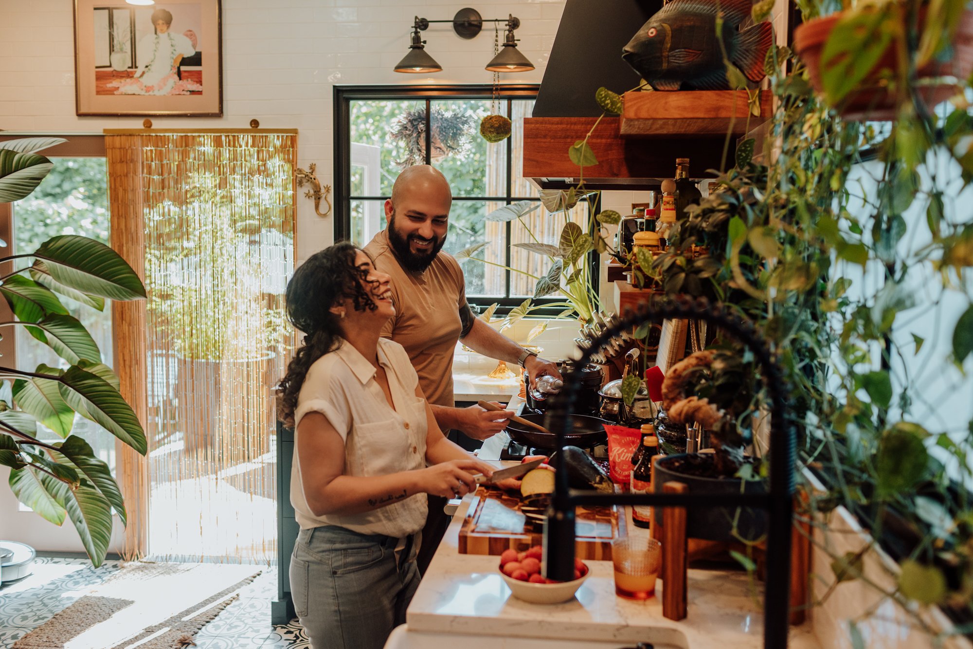Two people cooking in a cozy, plant-filled kitchen, smiling and enjoying each other's company.