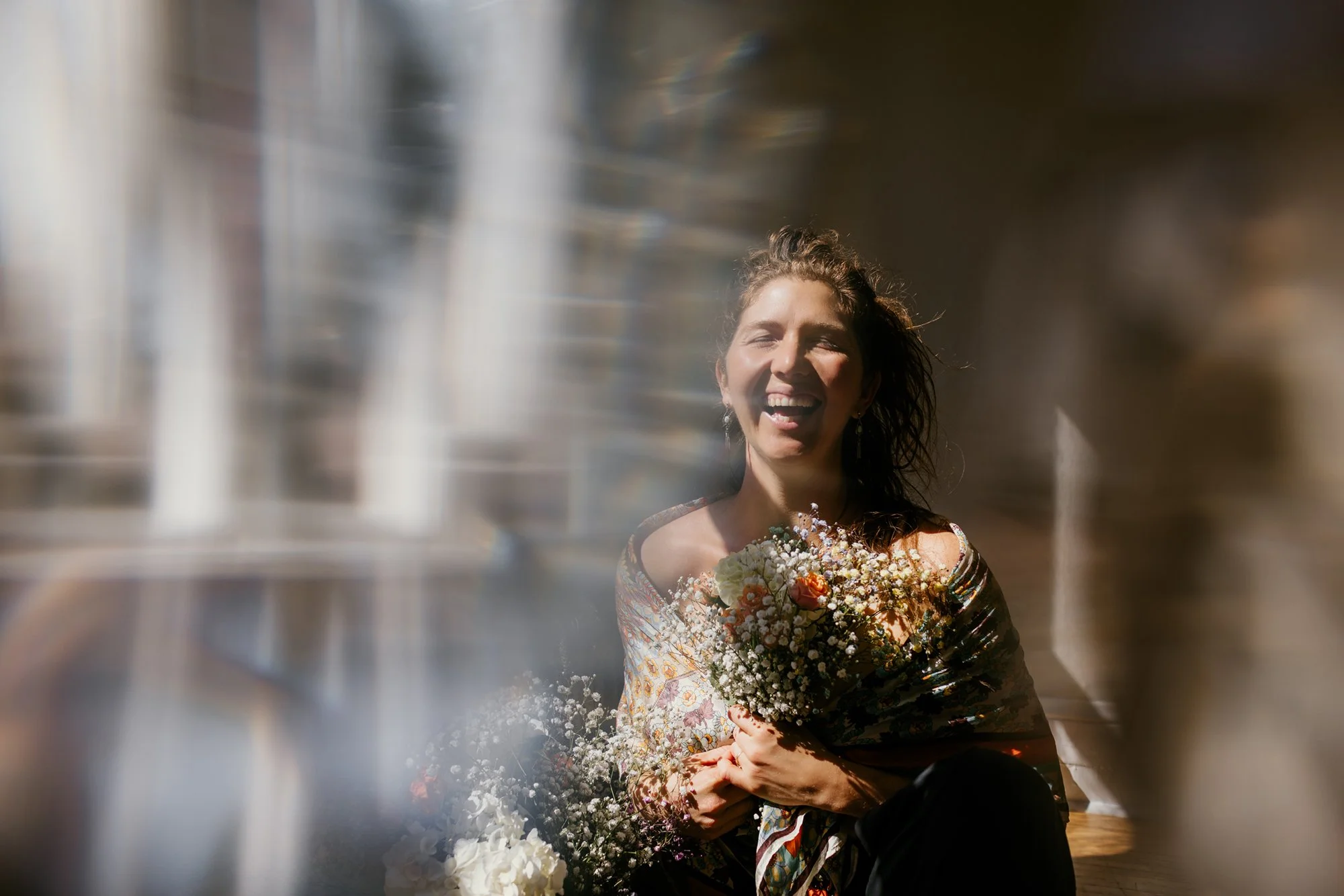 A woman smiling and holding a bouquet of flowers, sitting near a window with sunlight streaming in.