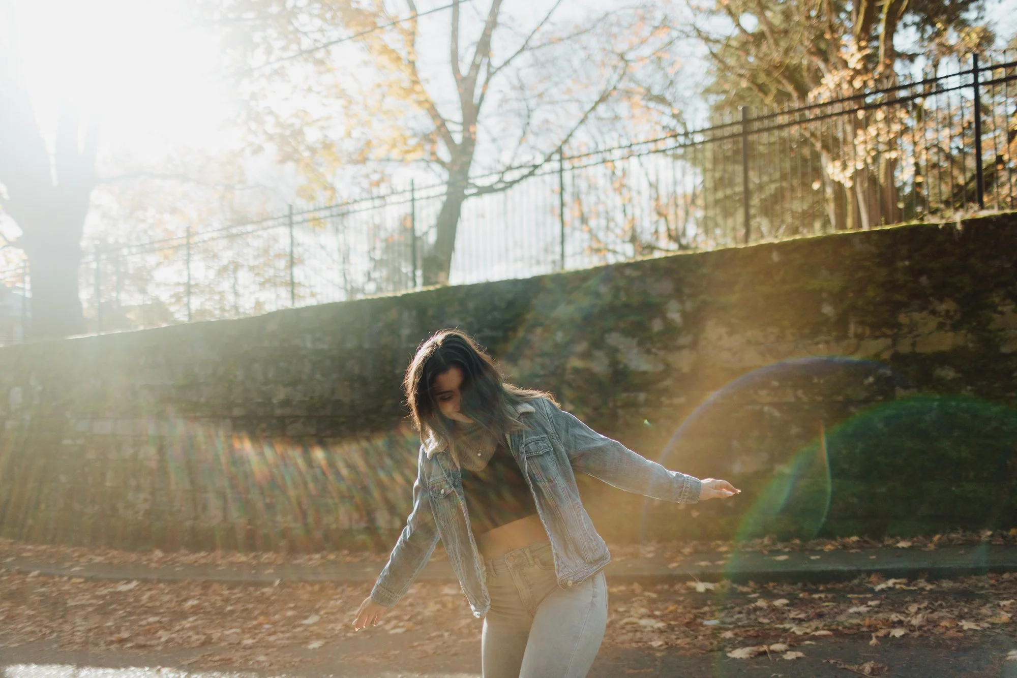 A young woman wearing a denim jacket and light pants walking outdoors on a sunny autumn day with fallen leaves, trees, and a fence in the background.