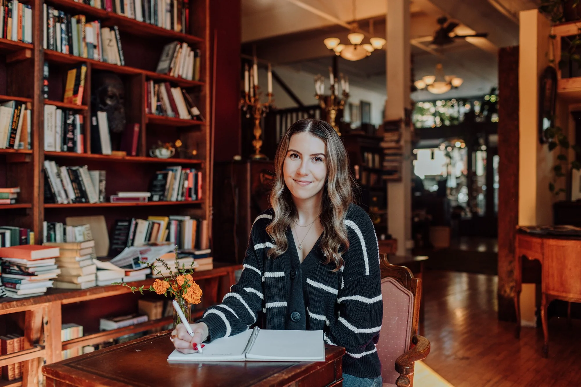 A young woman with wavy brown hair sitting at a wooden table, holding a pen in her right hand over an open notebook. She is smiling and wearing a black sweater with white stripes in a cozy, vintage-style bookstore or library with shelves of books and