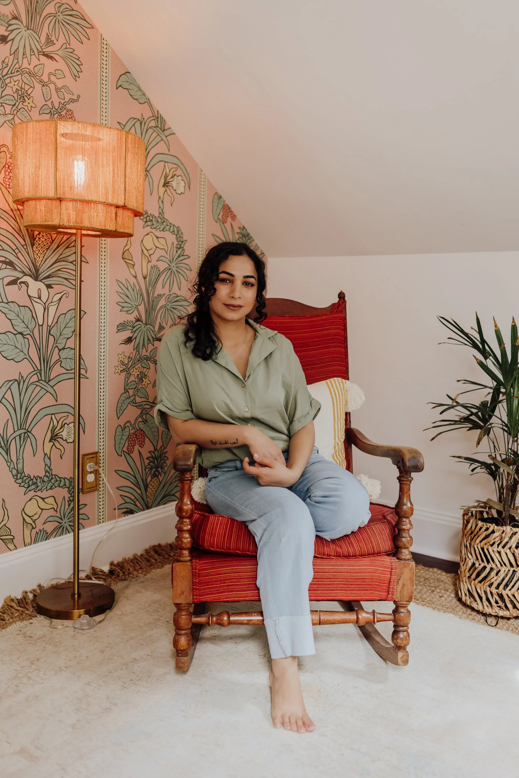 A woman with dark curly hair and light makeup, sitting barefoot on a vintage wooden armchair with red striped upholstery. She is wearing an oversized light green shirt and light blue jeans, in a cozy room with floral wallpaper, a floor lamp, a leafy 