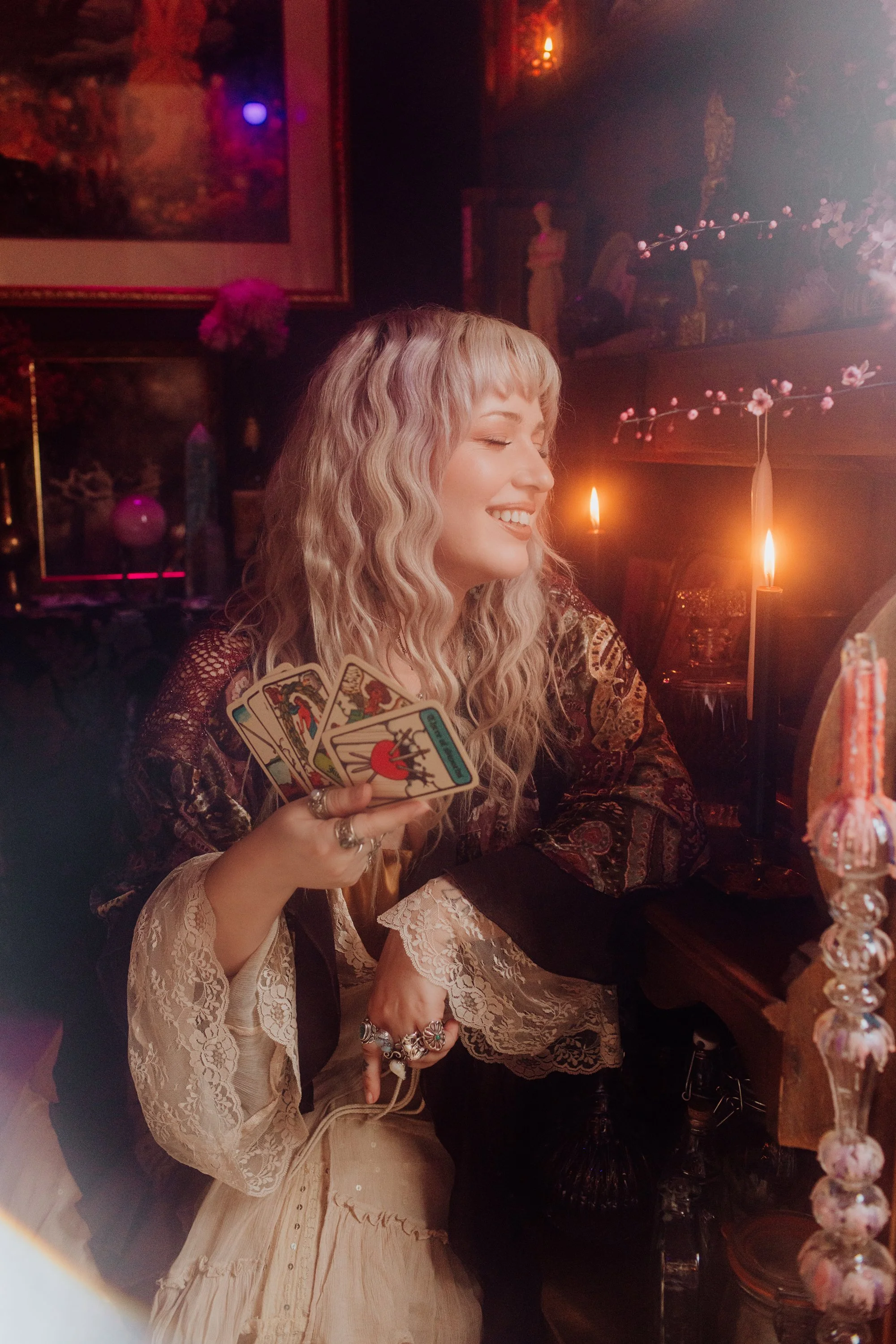 A woman with wavy blonde hair is smiling with her eyes closed, holding a fan of tarot cards. She is sitting in a dimly lit, cozy setting with candles and floral decorations around her.