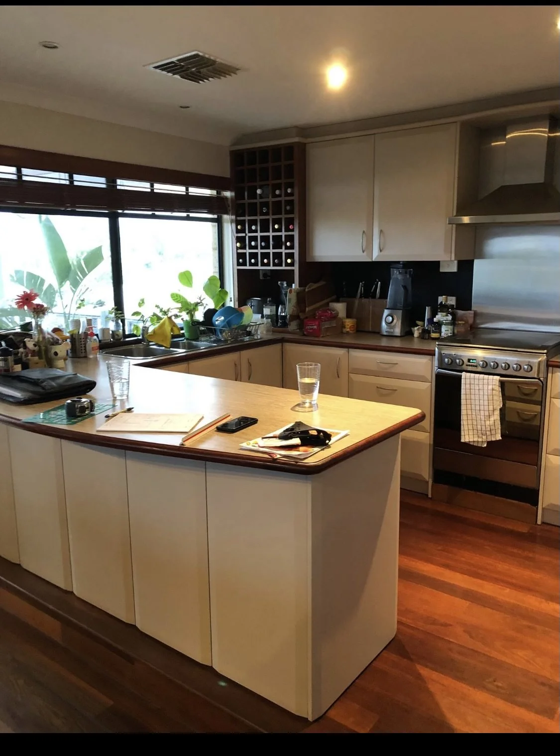 A modern kitchen with wooden cabinets and a kitchen island. The window has a view of green plants outside, and the counter has various kitchen items and potted plants. There are chairs, a stove, and a range hood.