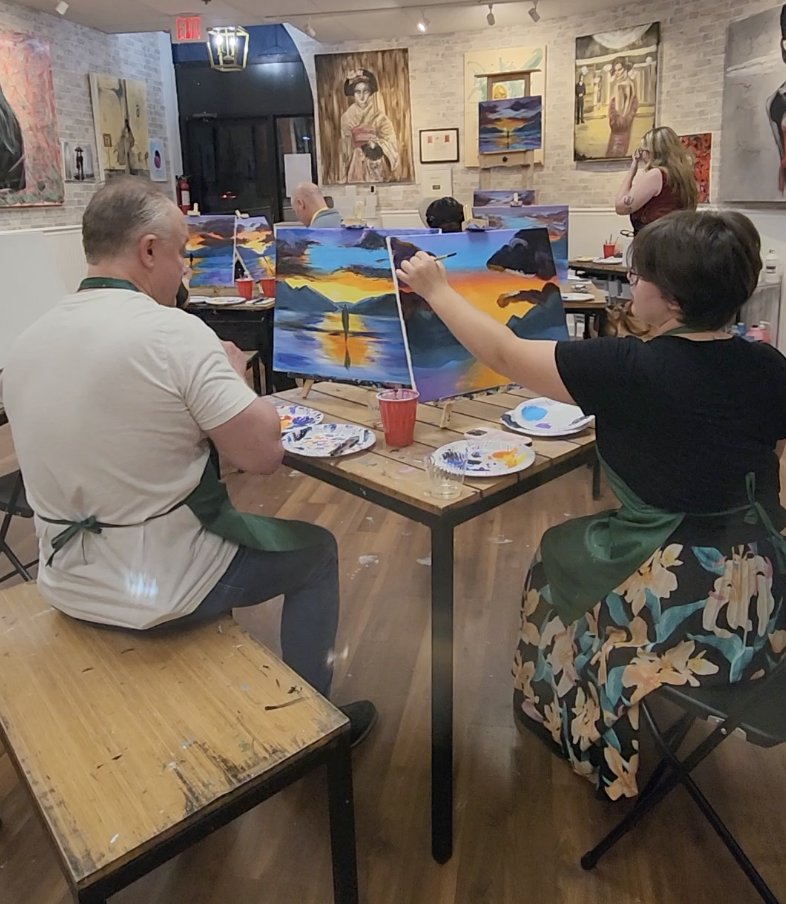 People participating in a paint and sip class, painting a sunset over water on their canvases in a cozy art studio.