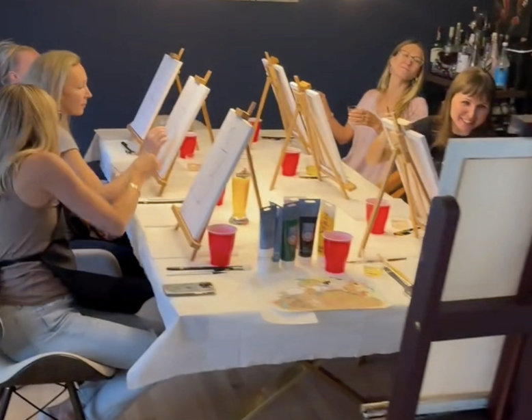 A group of women participating in a paint-and-sip event, sitting at a table with easels, paint, and drinks, in a social setting.