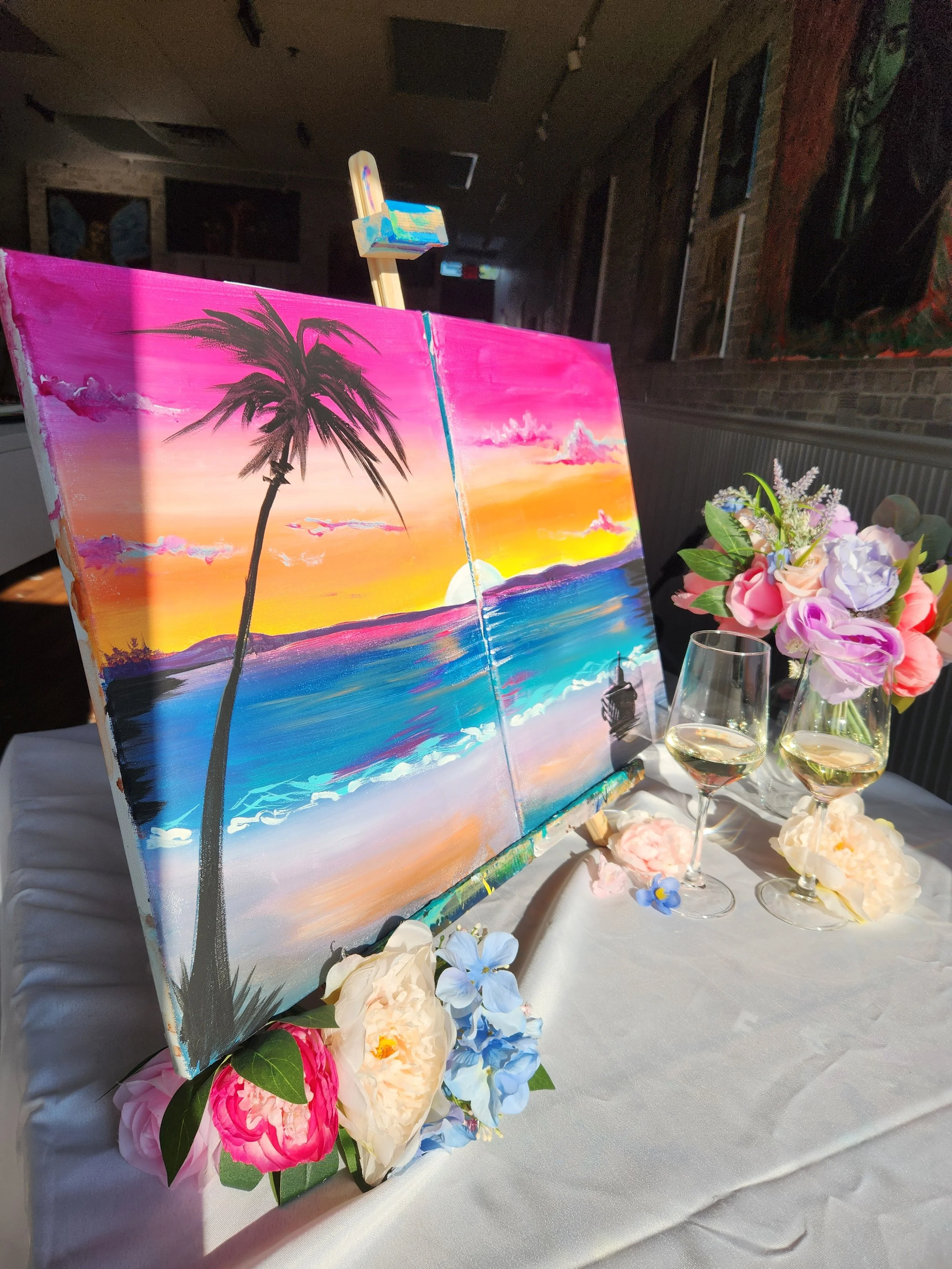 A colorful painting of a sunset over the ocean with a palm tree, displayed on an easel with pink, white, and blue flowers and two glasses of white wine on a white tablecloth.