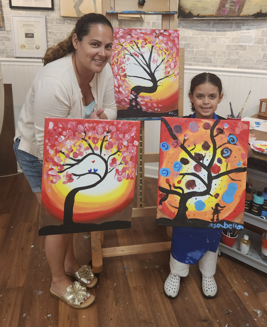 A woman and a young girl are standing in an art studio, each holding a colorful painting of a tree with a sunset background. The woman is smiling and has curly hair, while the girl is also smiling and has dark hair tied back. There is a larger painting of a tree with pink leaves and a sunset in the background on an easel behind them.