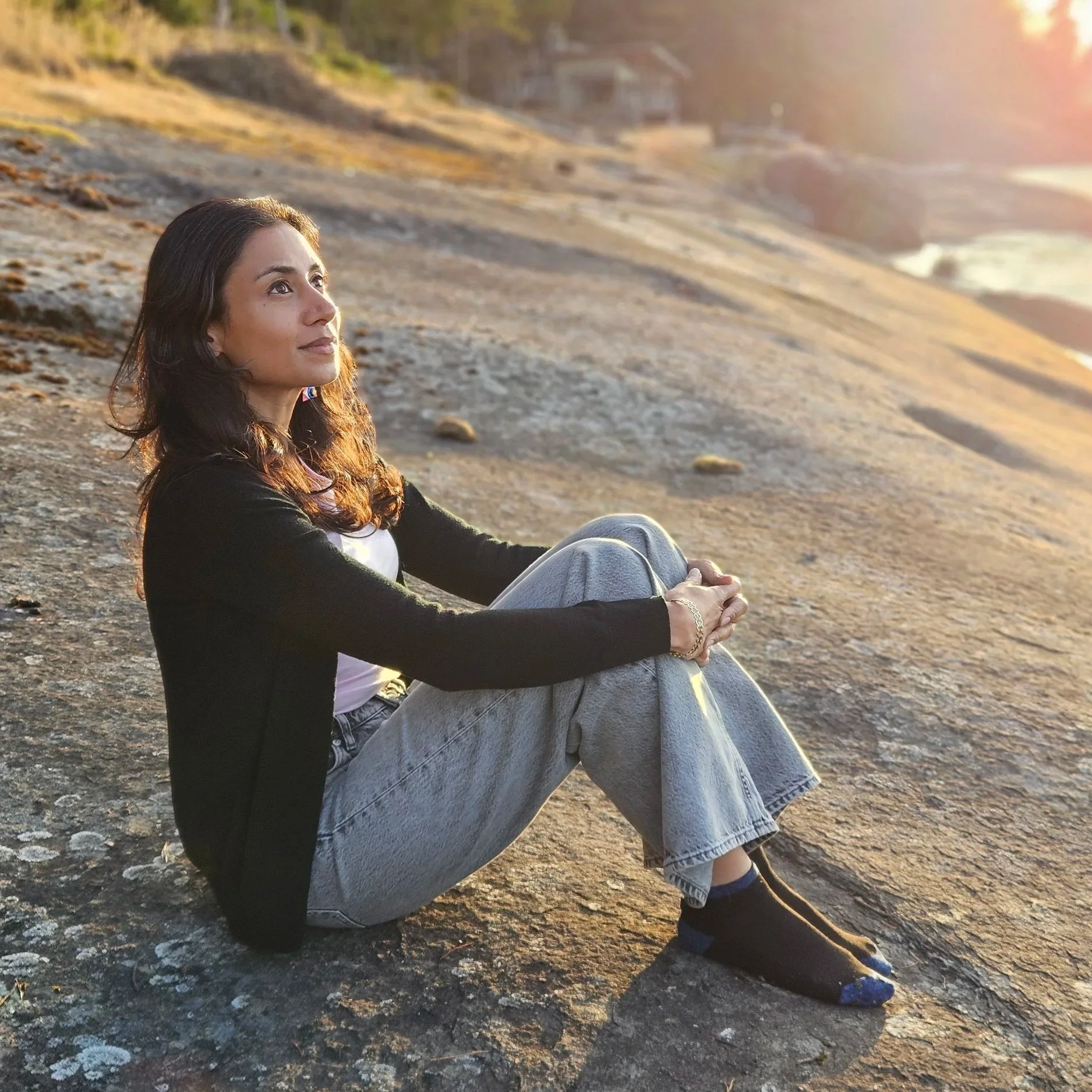 Preeti Kaur Dhaliwal sitting outdoors by the beach side with a black cardigan, pink top, and denim jeans
