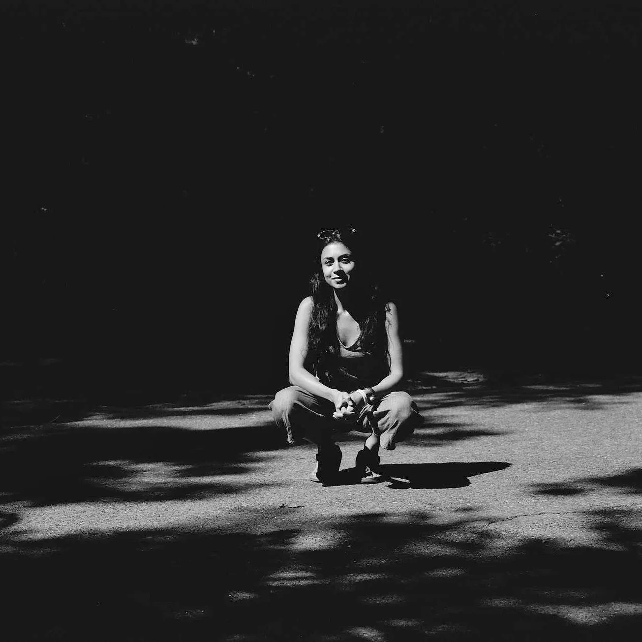 Preeti Kaur Dhaliwal with long hair squatting on a road with dappled shadows from leaves above, in an outdoor setting at night or in low light, captured in black and white.