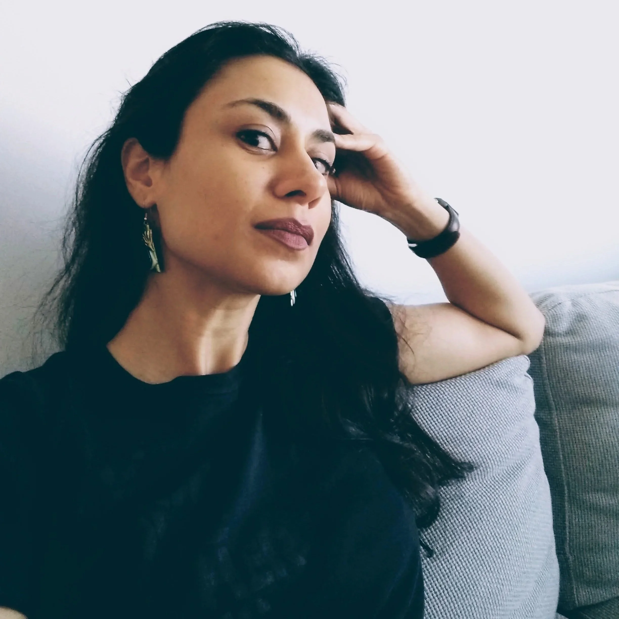 Preeti Kaur Dhaliwal with long dark hair and earrings sitting on a gray couch, posing with her head resting on her hand, wearing a black top and dark lipstick.