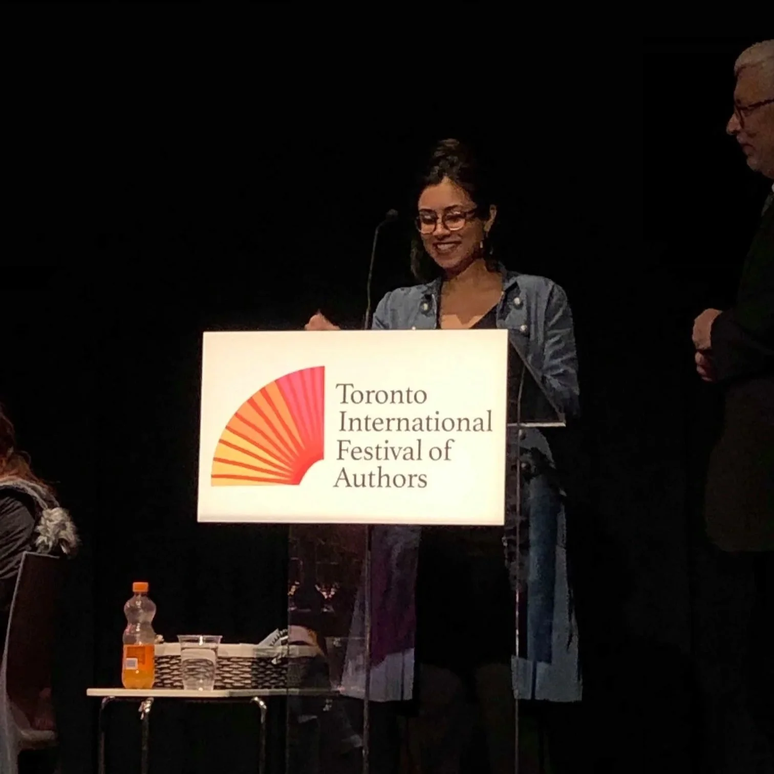 BIPOC artist standing at a podium with a sign reading 'Toronto International Festival of Authors'. She is smiling and wearing glasses and a denim jacket.