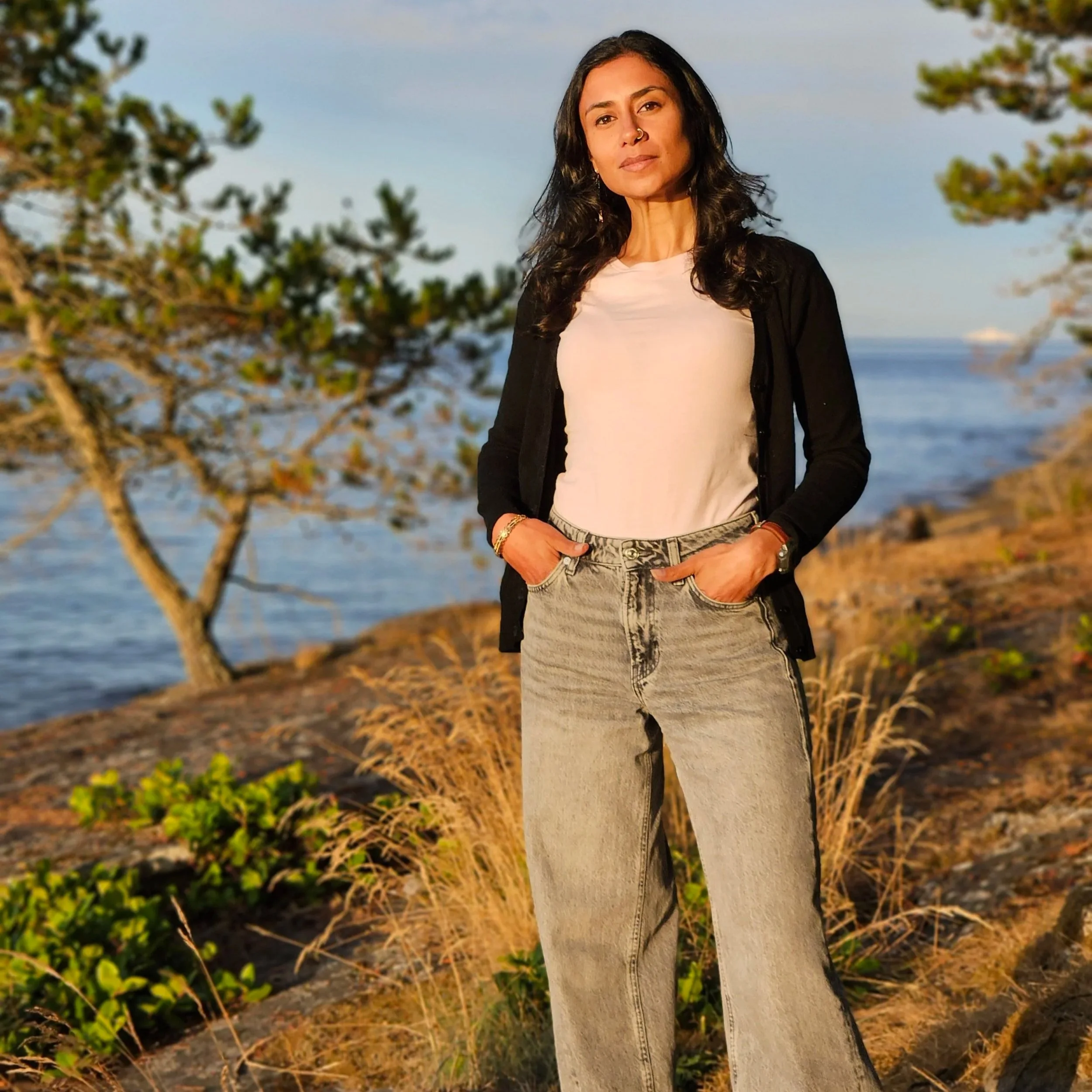 BIPOC artist/brown-skinned woman standing outdoors by the ocean during golden hour, wearing a light pink top, black cardigan, and denim jeans