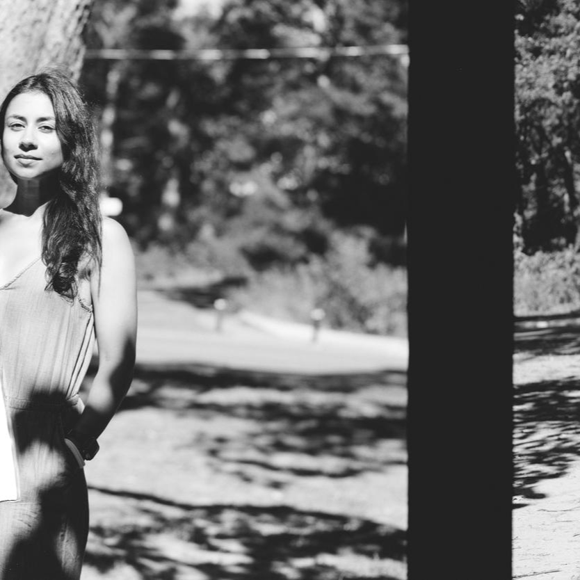 A black and white photo of a Preeti Dhaliwal with long, curly hair standing outdoors next to a tree, with a blurred background of trees and a winding path.