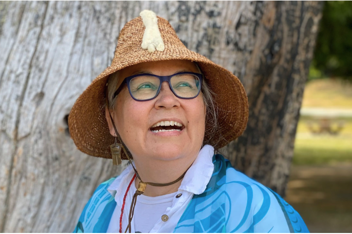 A woman with glasses wearing a straw hat with a small animal figure on it, smiling outdoors near a tree.