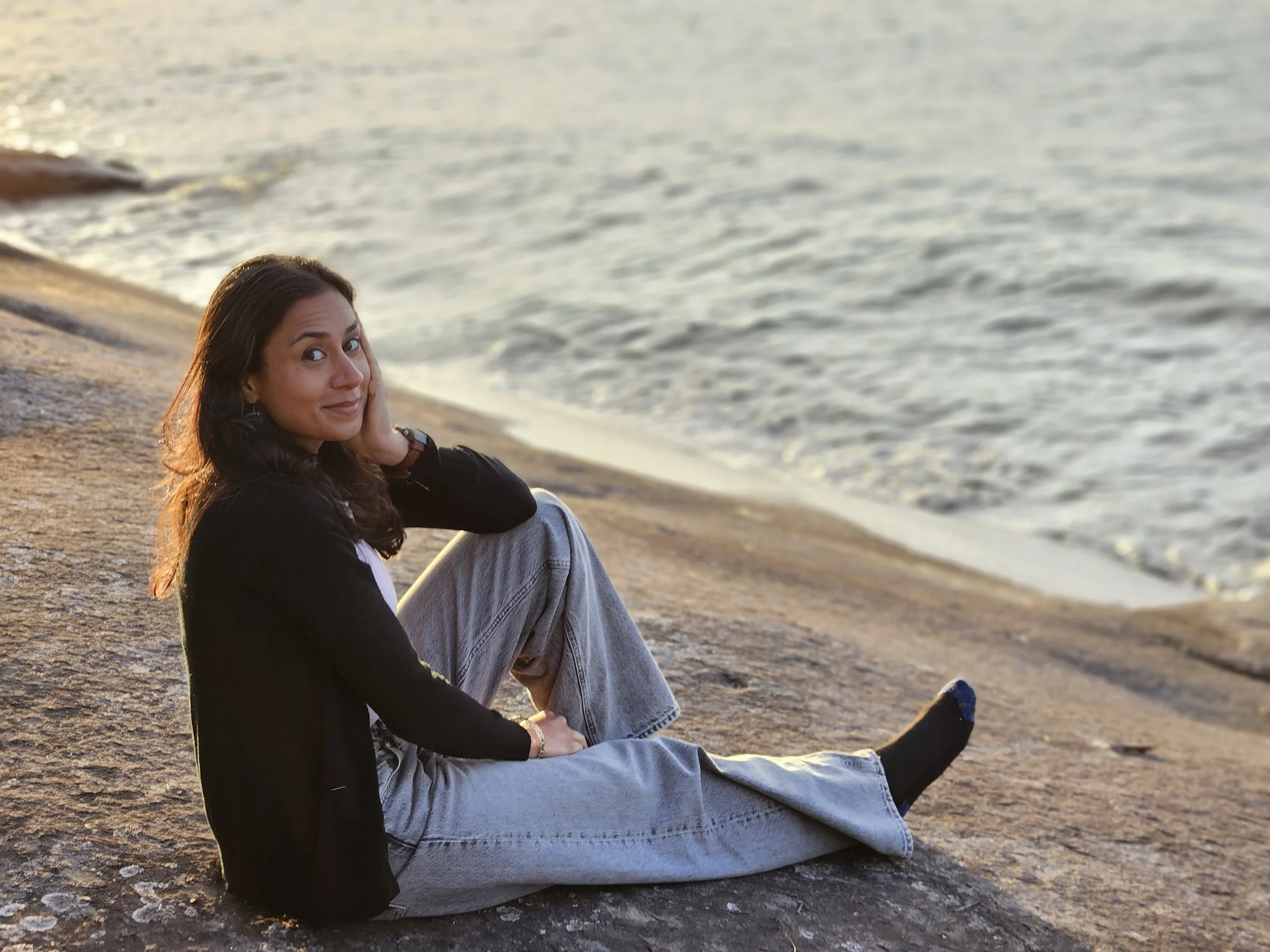 Preeti Kaur Dhaliwal sitting by ocean side with a black cardigan and denim jeans