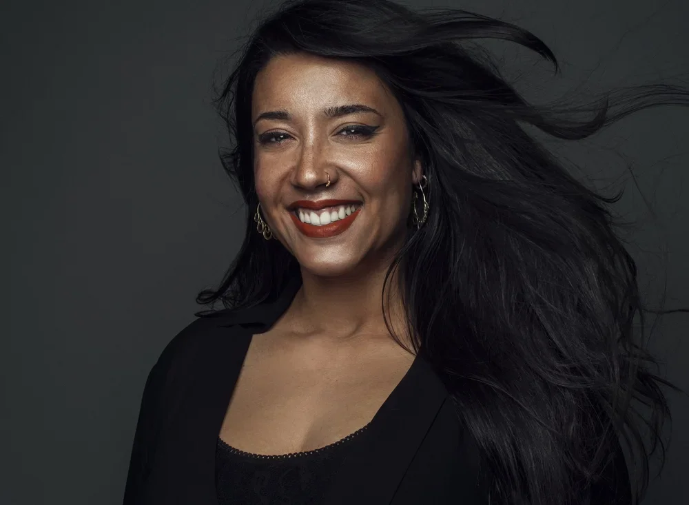 Smiling woman with dark hair blowing to the side, wearing earrings and a black top, against a dark background.