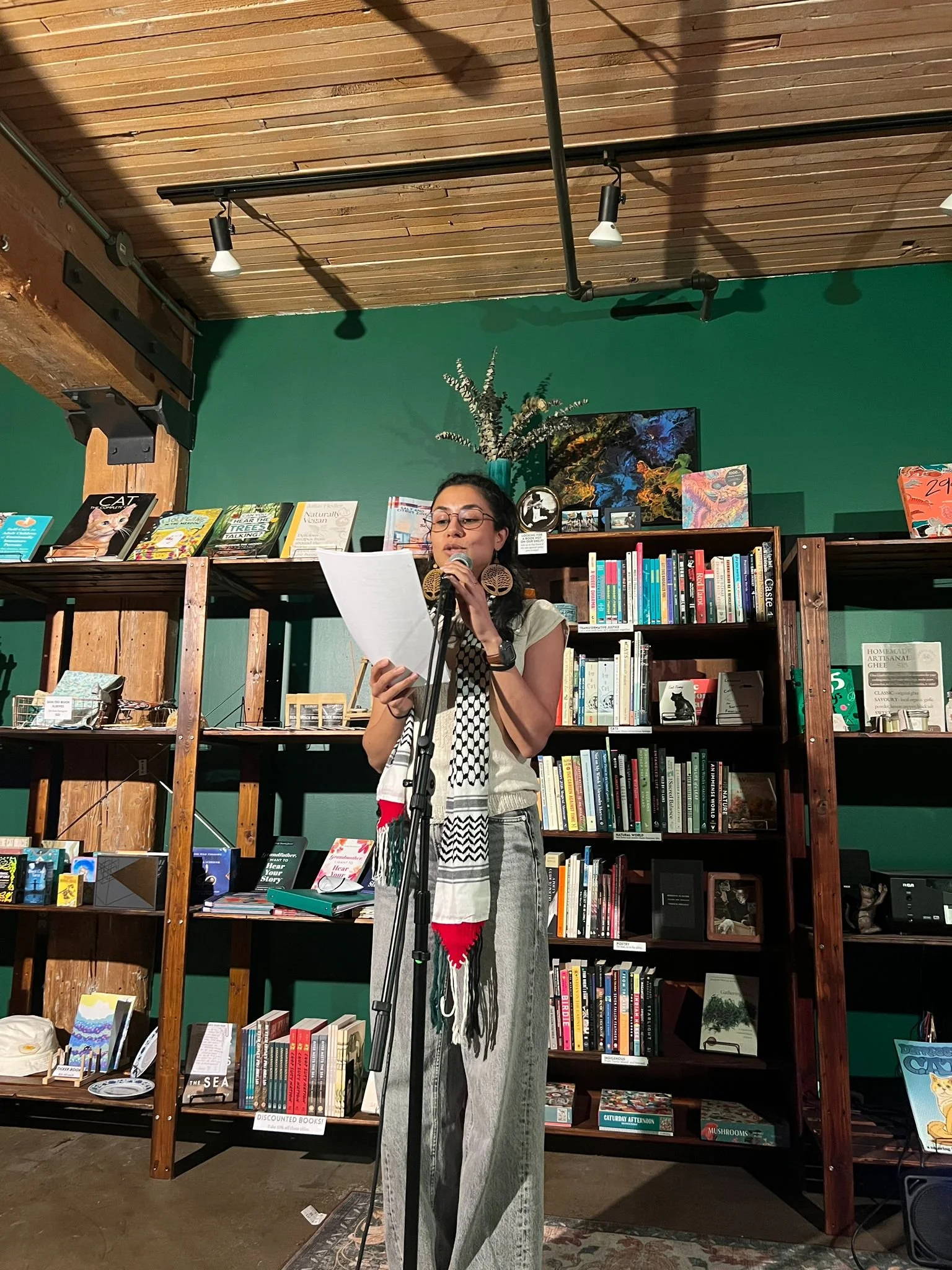 A woman stands in a bookstore or library, reading aloud into a microphone while holding a paper. She wears glasses, large earrings, a scarf, and casual clothing. Behind her are wooden bookshelves filled with books and decorative items, with a green wall and a ceiling with wooden panels.