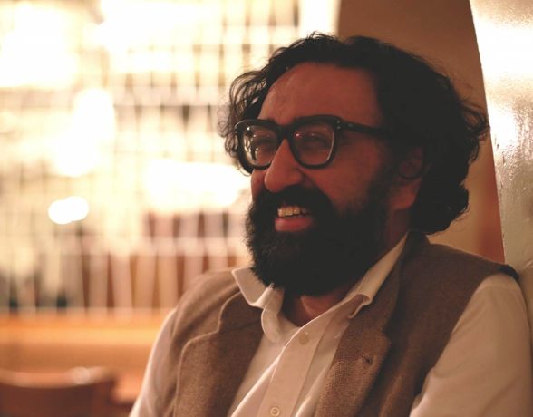 A man with glasses, a beard, and curly hair smiling while sitting indoors in a warmly lit environment.