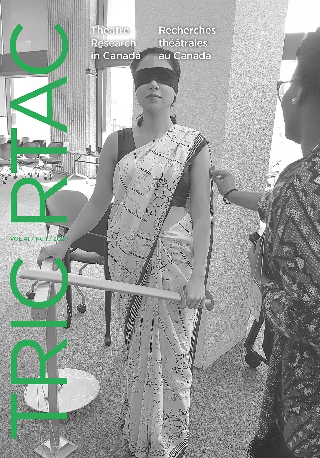 A woman wearing a traditional sari blindfolded and holding a cane, is being guided by another woman in an indoor setting. The scene appears to be part of a research or educational activity related to theatrical research in Canada.