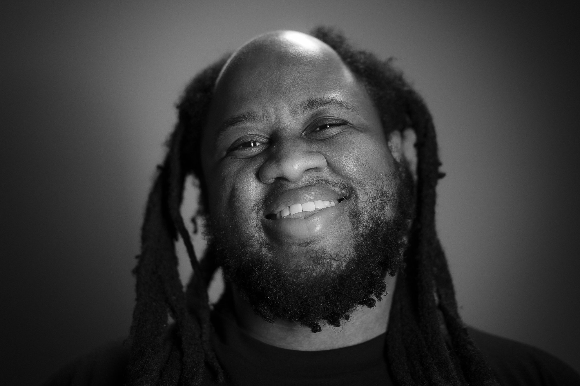 Black and white portrait of a smiling man with dreadlocks and a beard, facing the camera.