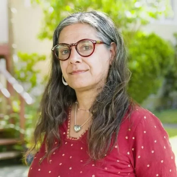 A woman with long gray hair, glasses, and hoop earrings, wearing a red top with gold accents, standing outdoors with green trees in the background.