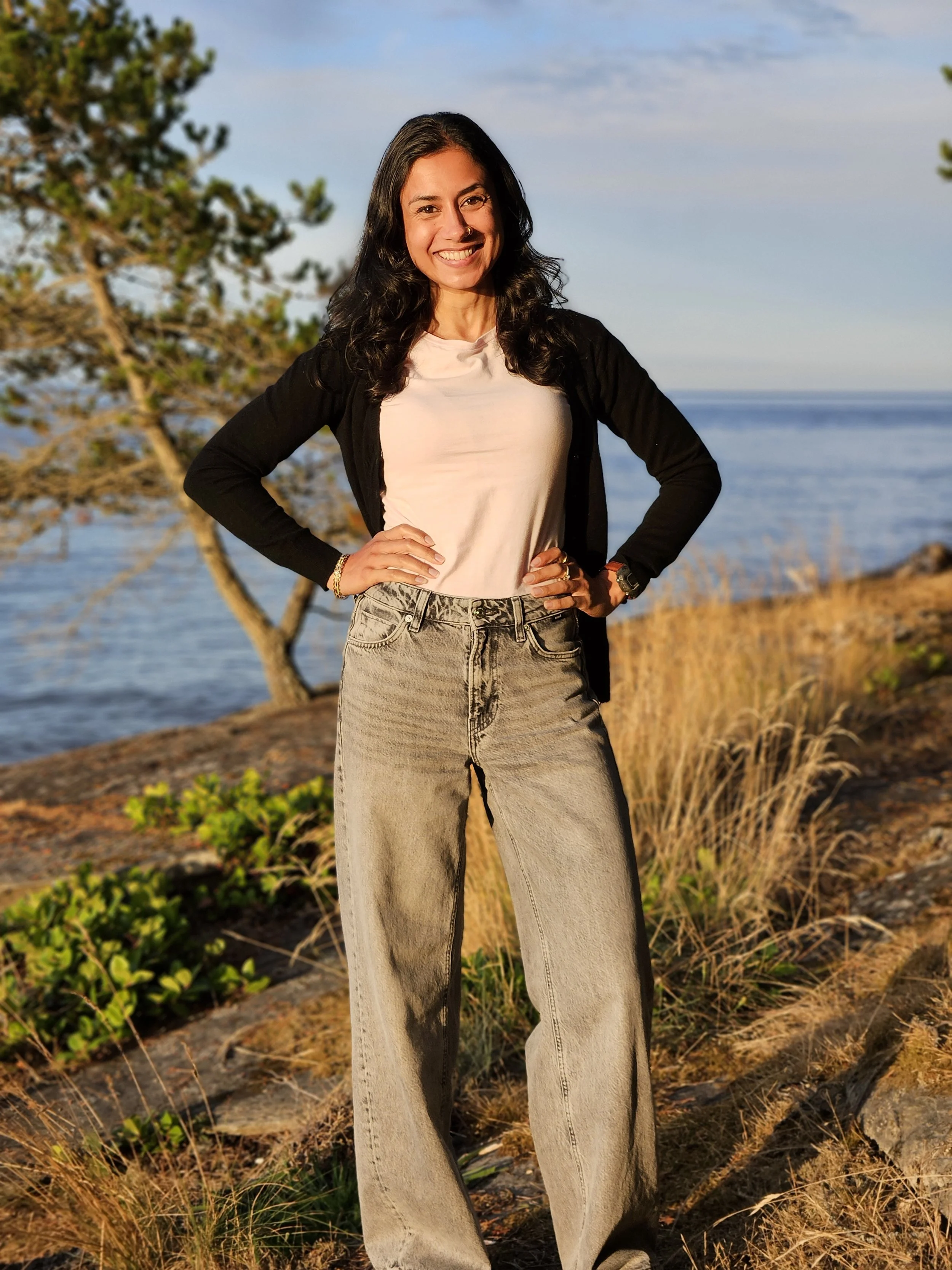 Preeti Kaur Dhaliwal standing outdoors in golden hour with a smile on her face wearing a black cardigan, pink shirt, and denim jeans