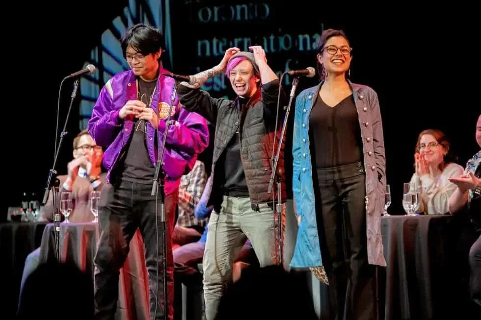 People standing on stage during an event, with some clapping and smiling, while others are sitting at tables with drinks, and a large screen displaying text in the background.
