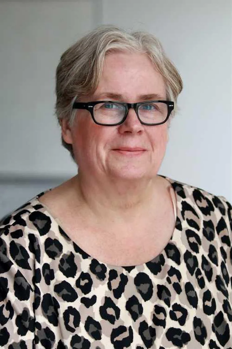 An older woman with short gray hair, black glasses, wearing a leopard print blouse, smiling gently in an indoor setting.