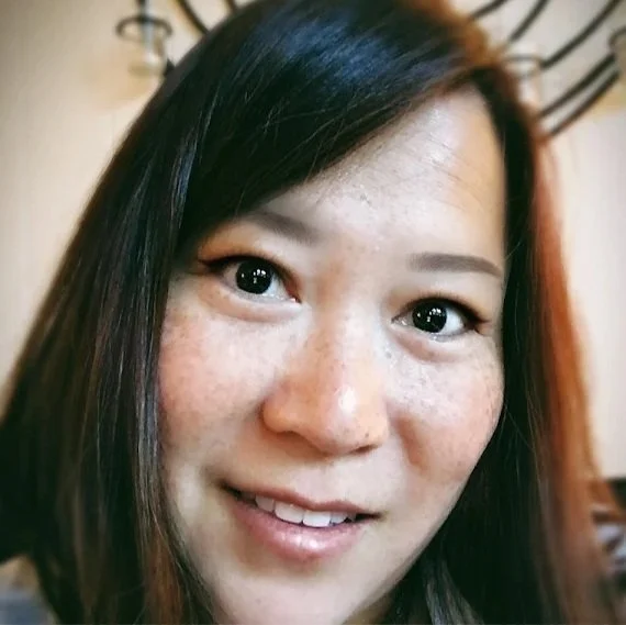 Close-up of a smiling woman with long dark hair and freckles in an indoor setting.