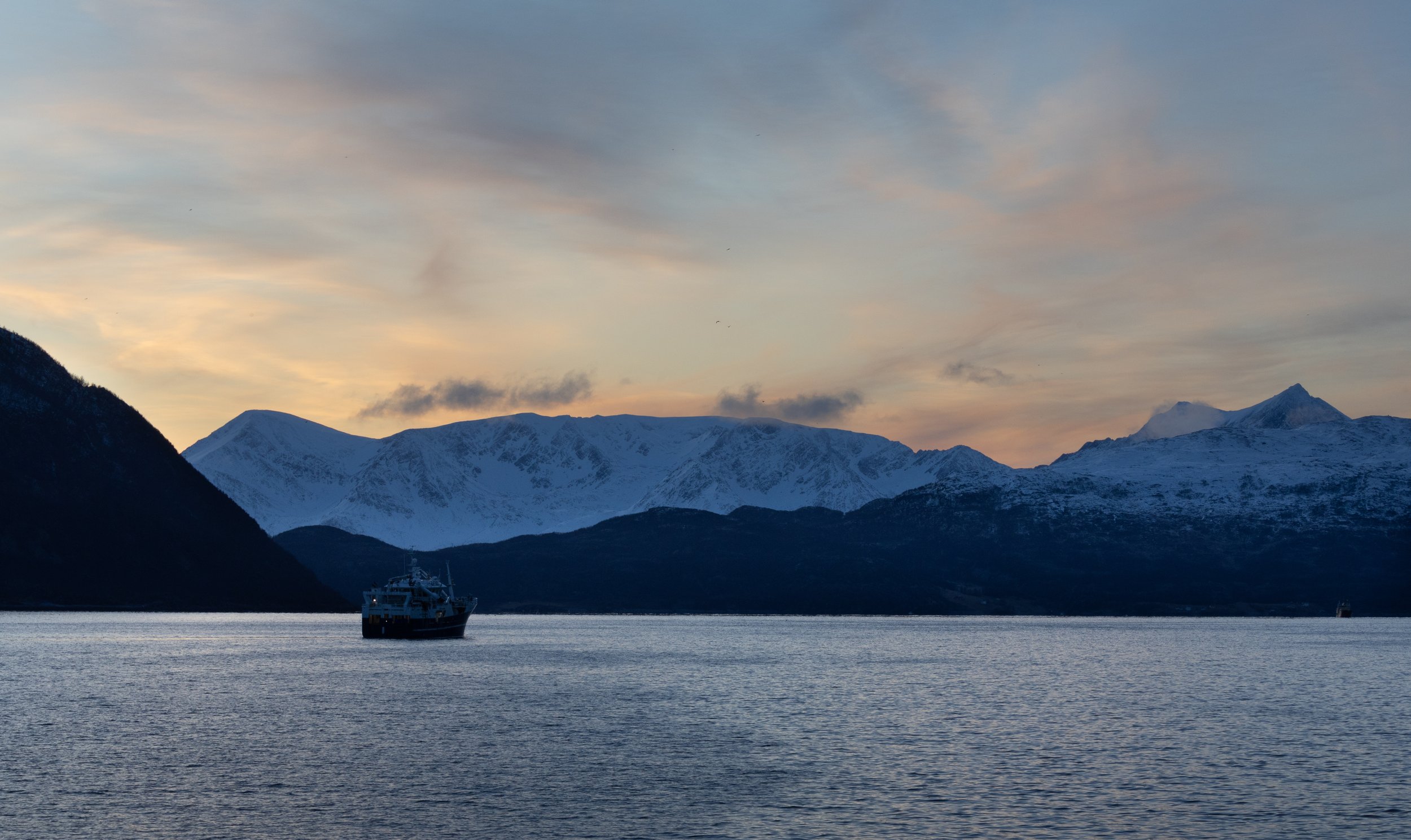 Fishing boat on a calm sea with snow-covered mountains and a sunset sky in the background.