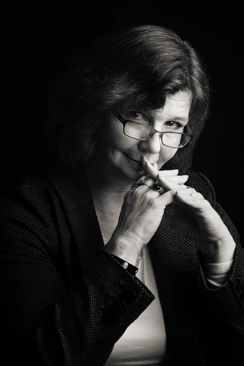 A black and white portrait of a woman with glasses, looking mischievously at the camera, fingers touching her lips, wearing a dark textured blazer and jewelry.